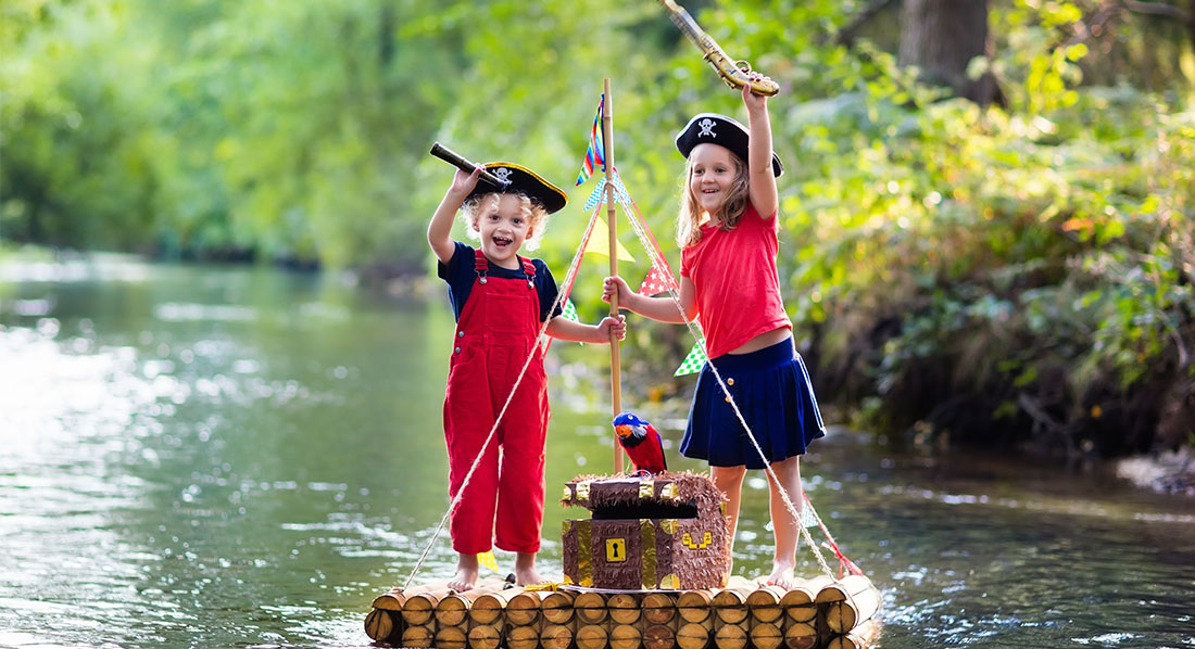 Zwei Kinder stehen freudig auf einem Floß im Bach, zwischen ihnen eine Schatzkiste